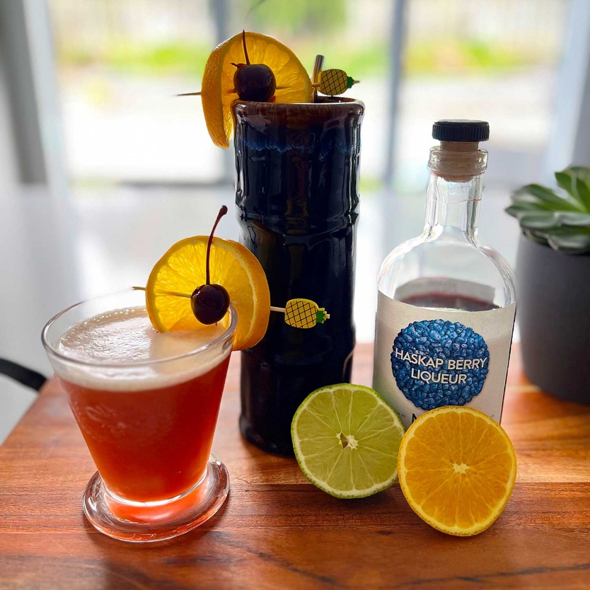 Haskap Berry cocktail on a counter with lemon, lime, and Haskap Berry liqueur. Haskap Sling on a counter with lemon, lime, and Haskap Berry liqueur.